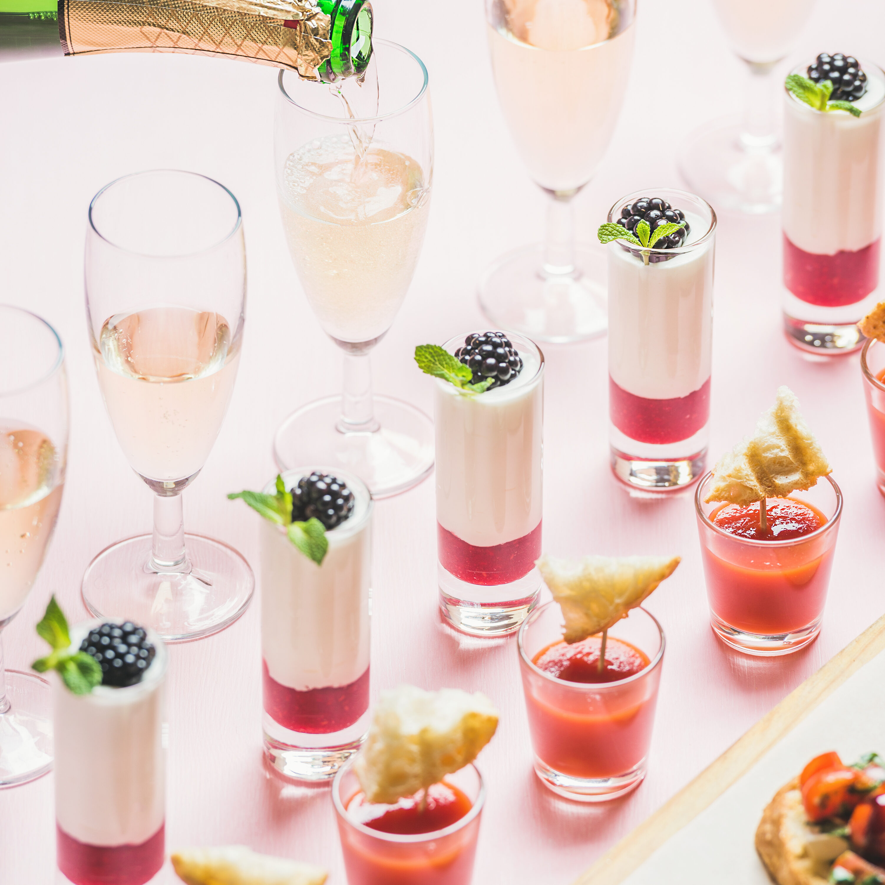 Various snacks, brushettas, gazpacho shots, desserts with berries and champagne pouring to glasses on corporate event, christmas, wedding celebration over pastel pink background, selective focus